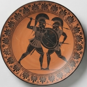 Assiette en terre cuite à décor noir : deux guerriers casqués au combat, frise de palmettes.